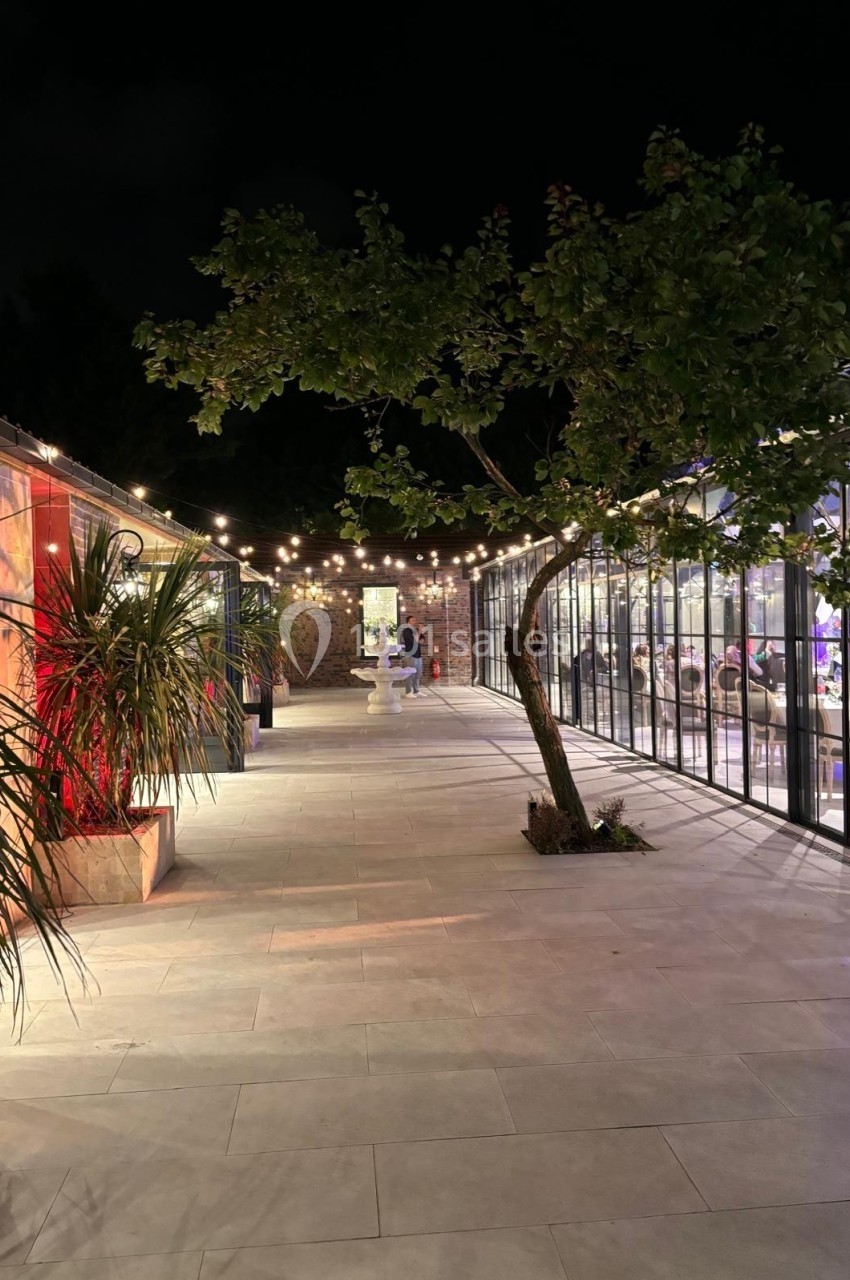 Location salle Bobigny (Seine-Saint-Denis) - Orangerie du Manoir des Vignes #10 Allée extérieure éclairée la nuit, bordée de plantes, d'un arbre et de bâtiments vitrés illuminés.