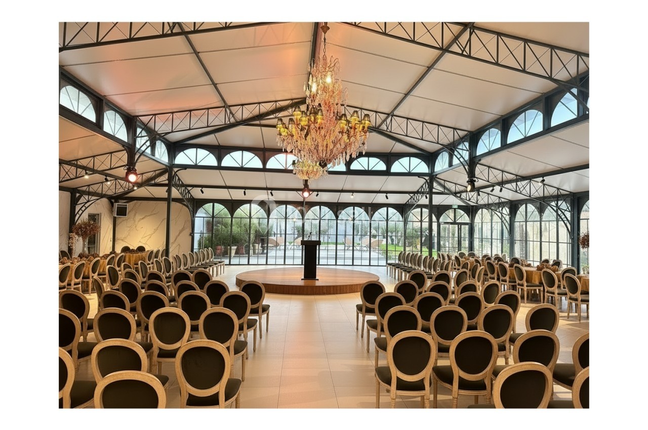 Location salle Bobigny (Seine-Saint-Denis) - Orangerie du Manoir des Vignes #5 Salle de réception lumineuse avec des rangées de chaises disposées face à une estrade et un pupitre sous un grand lustre.
