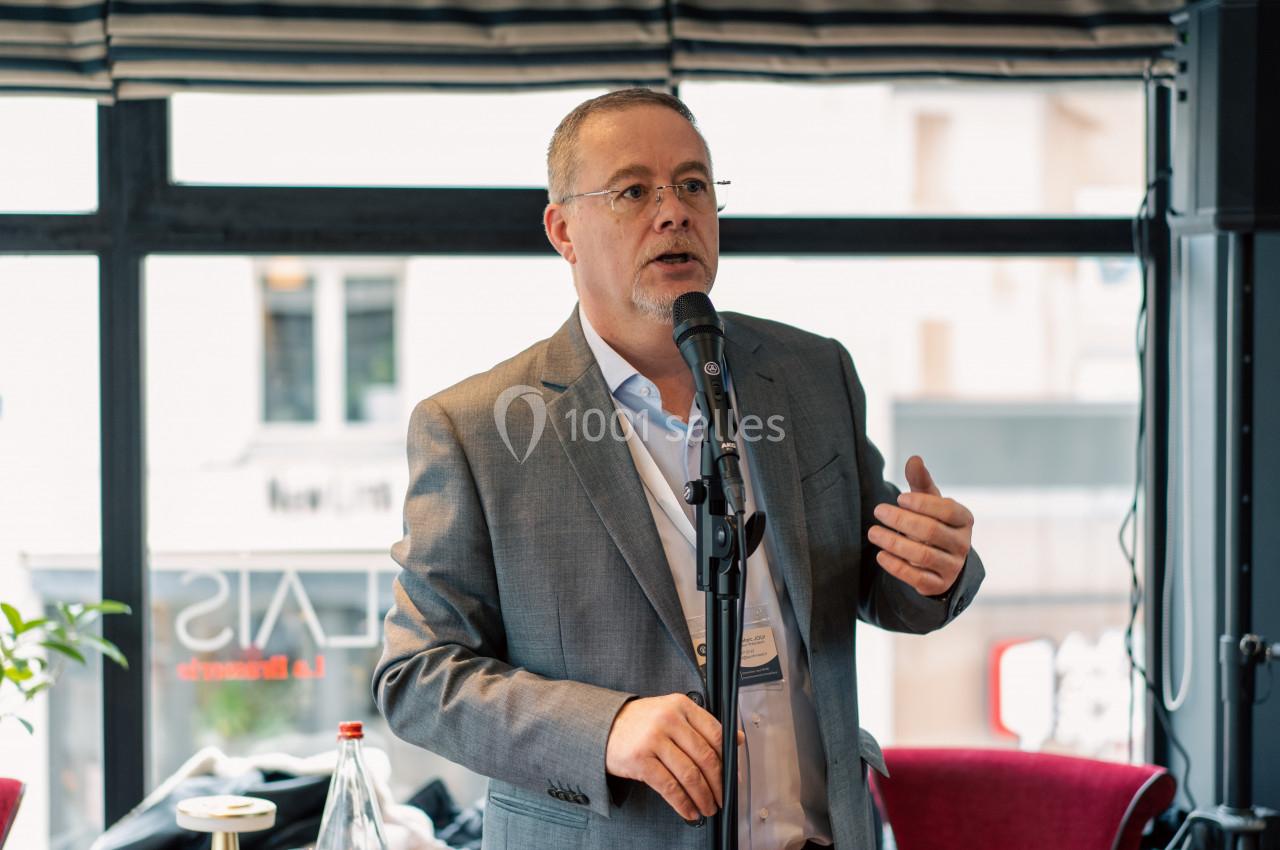 Un homme en costume parle dans un micro, debout dans une salle lumineuse avec des fenêtres en arrière-plan.
