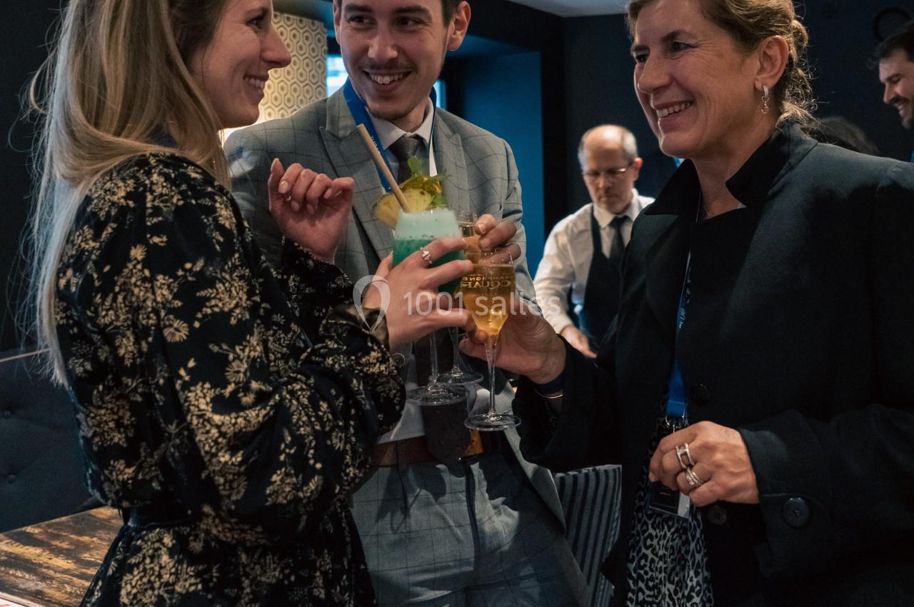 Trois personnes discutent et trinquent dans un cadre intérieur convivial, éclairé par des lumières tamisées.