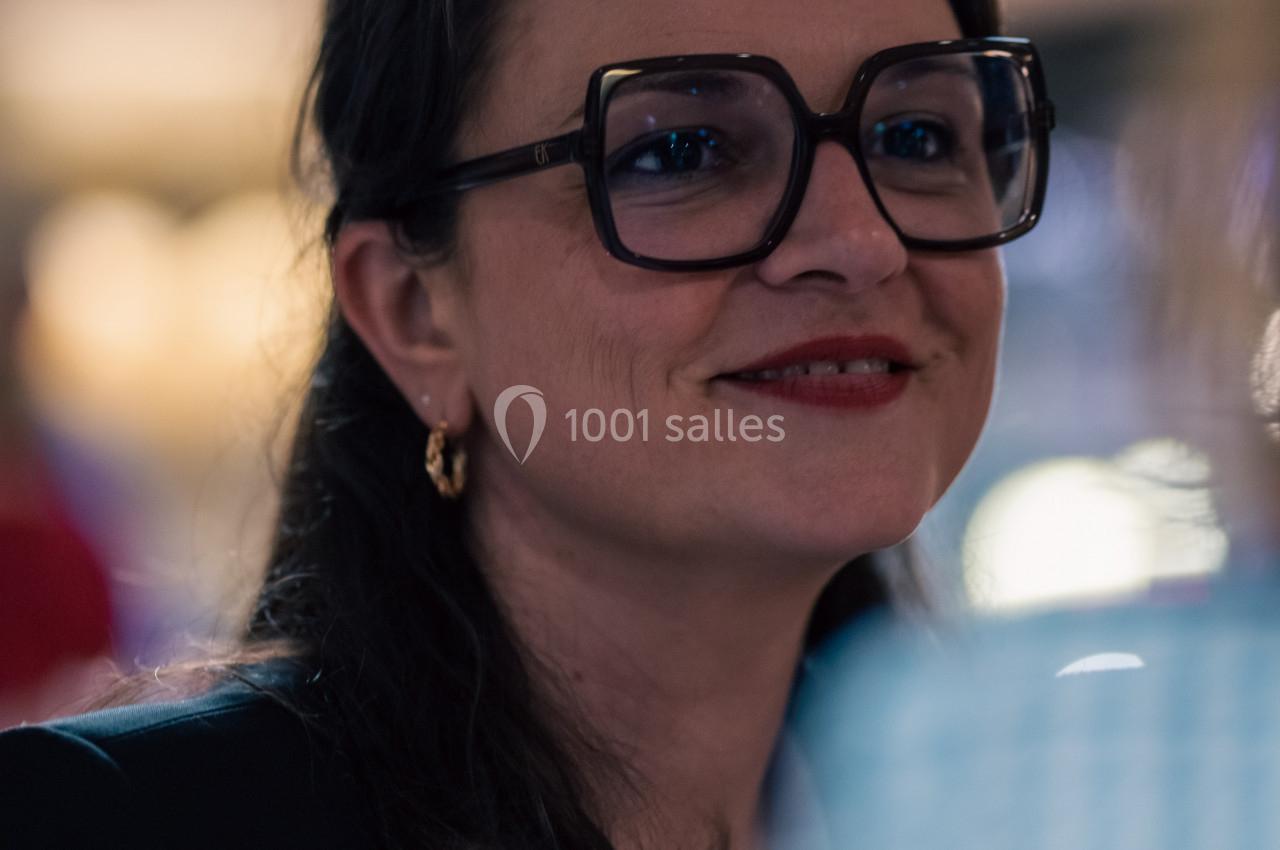 Femme portant des lunettes épaisses et un blazer, souriant dans un environnement intérieur flou.