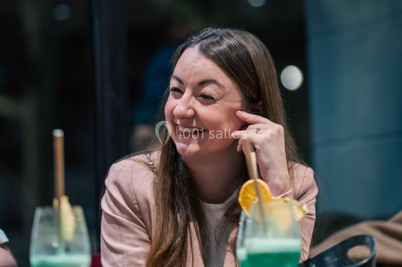 Une femme souriante assise à une table avec des cocktails verts et un plateau de nourriture devant elle.