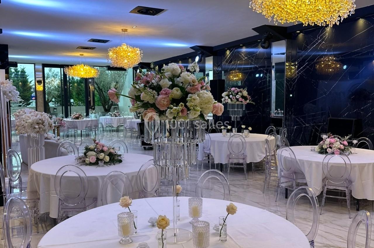 Salle de réception élégante avec tables rondes décorées de fleurs et chaises transparentes sous des lustres lumineux.