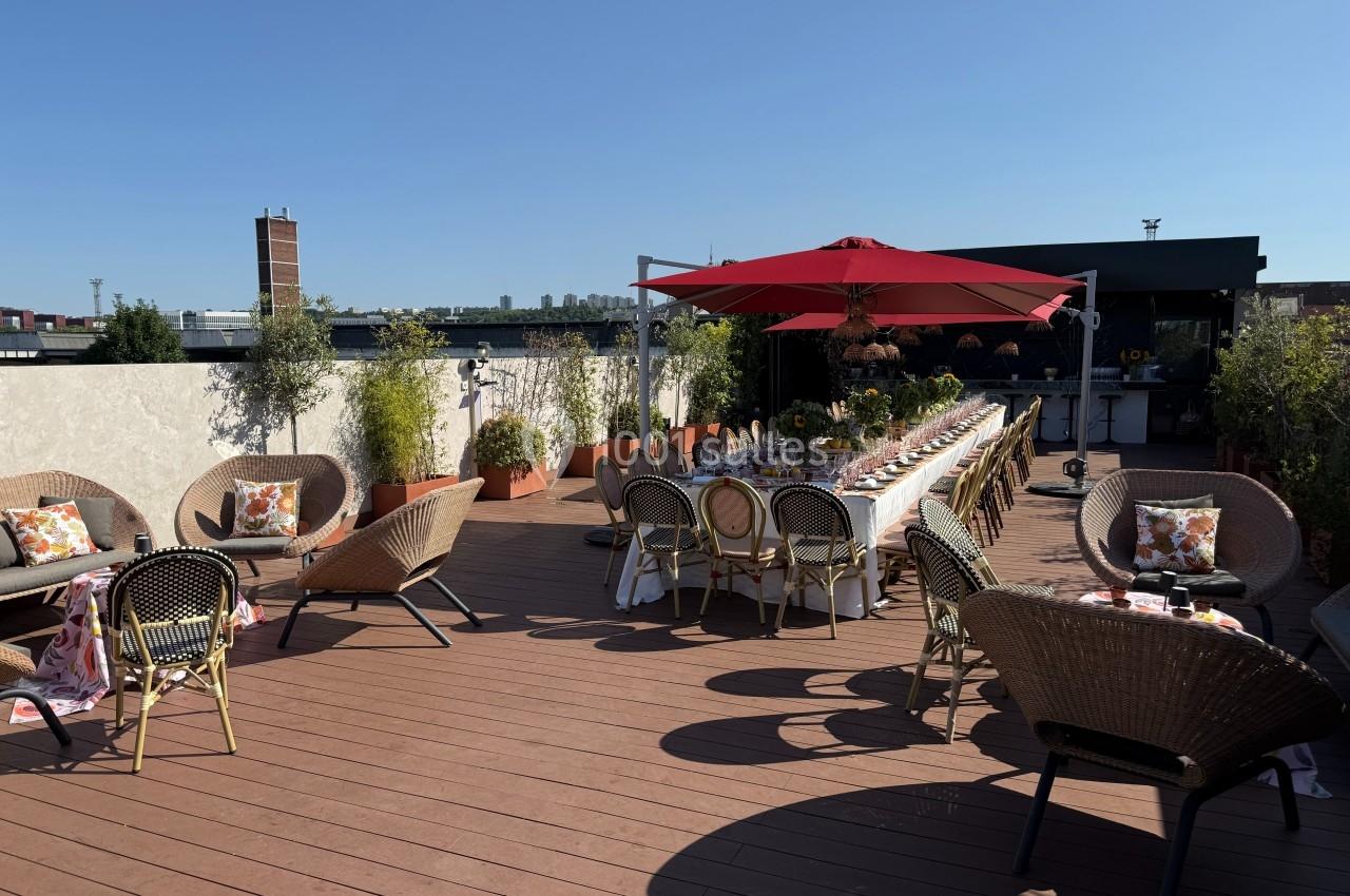 Terrasse aménagée avec tables dressées, chaises en rotin, parasol rouge et végétation en arrière-plan.