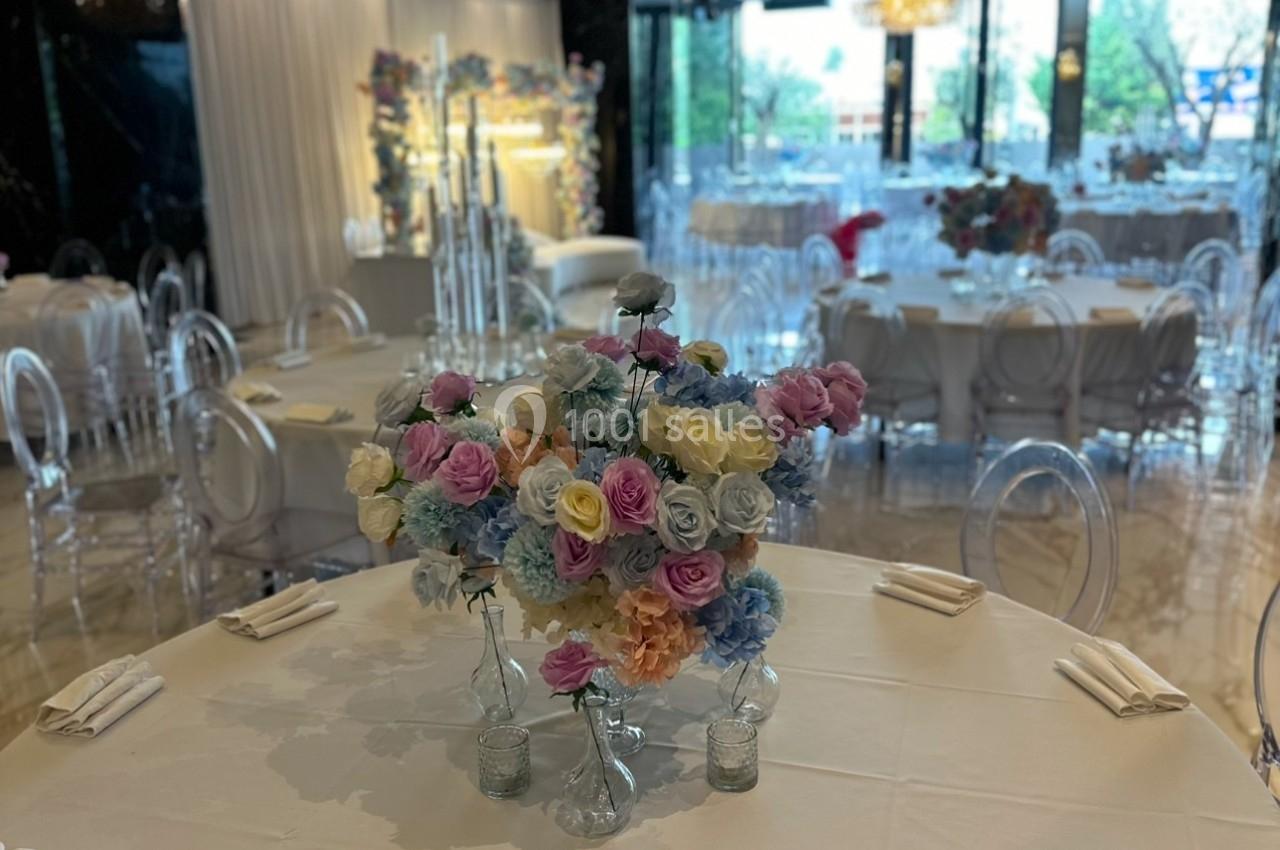 Salle de réception élégante avec tables rondes dressées, décorées de bouquets de fleurs colorées et chaises transparentes.