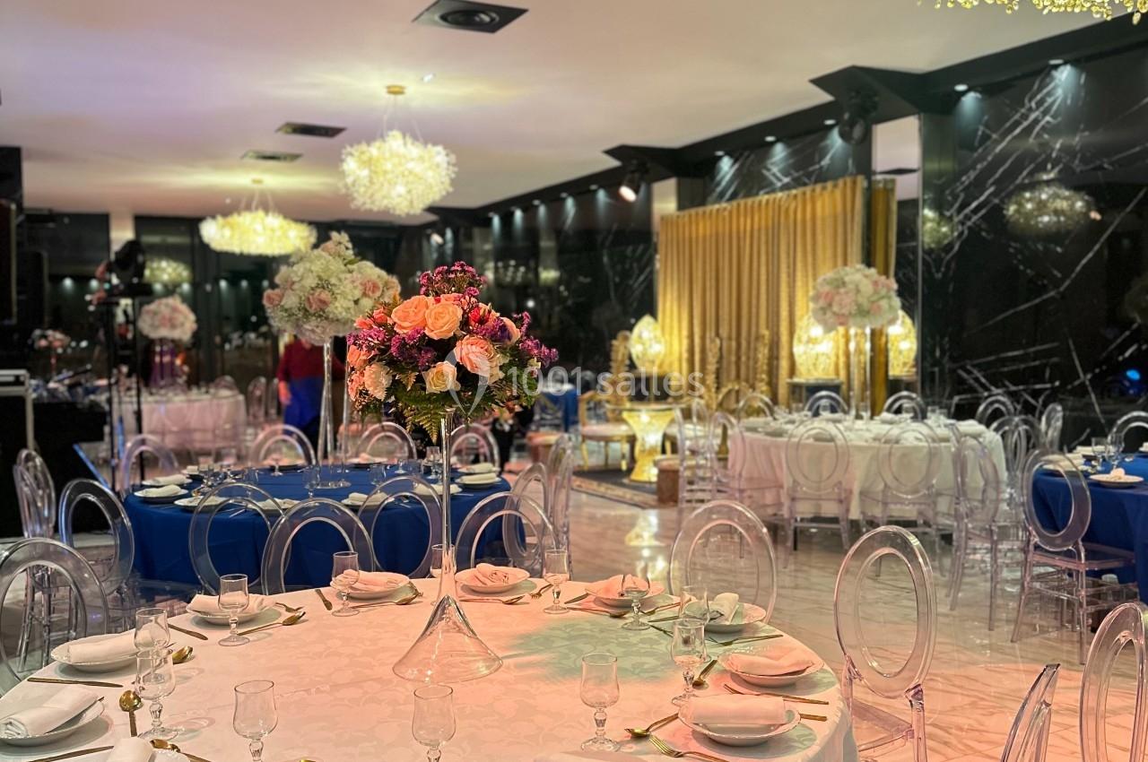 Salle de réception élégante avec tables dressées, chaises transparentes et décorations florales sous des lustres lumineux.