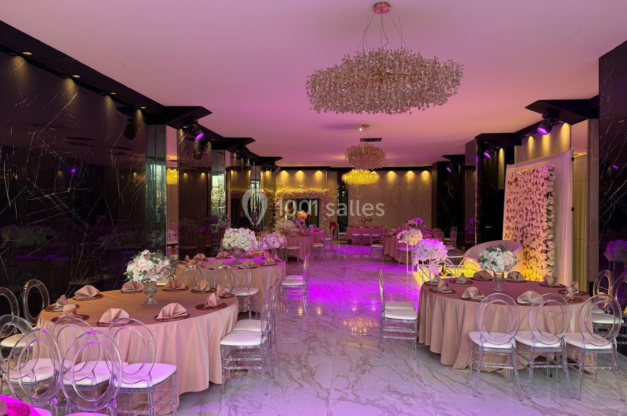 Salle de réception élégante avec tables rondes dressées, chaises transparentes et éclairage violet tamisé.