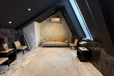 Salon mansardé avec canapé gris, fauteuils, tables d'appoint et éclairage naturel provenant d'une fenêtre inclinée.