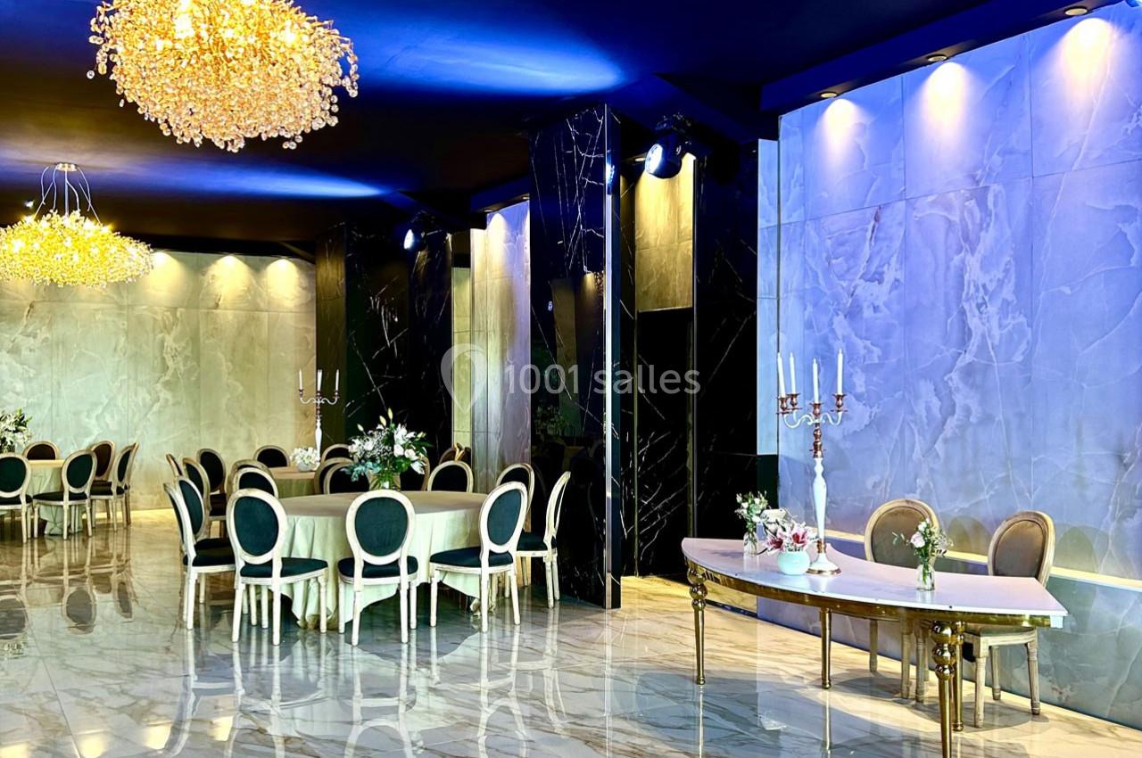 Salle de réception élégante avec tables rondes dressées, chandeliers et éclairage tamisé sur des murs en marbre.