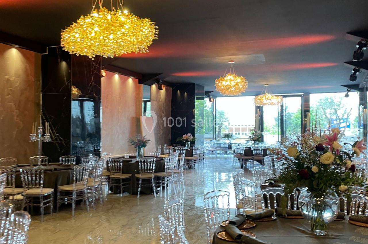 Salle de réception élégante avec tables dressées, chaises transparentes et lustres lumineux sous un plafond sombre.