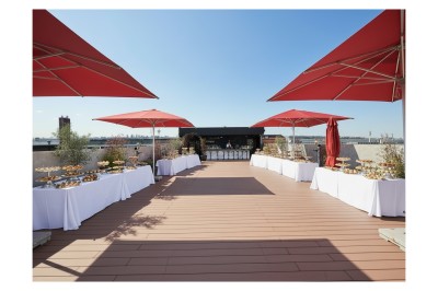 Terrasse ensoleillée aménagée pour un événement, avec tables dressées et parasols rouges.