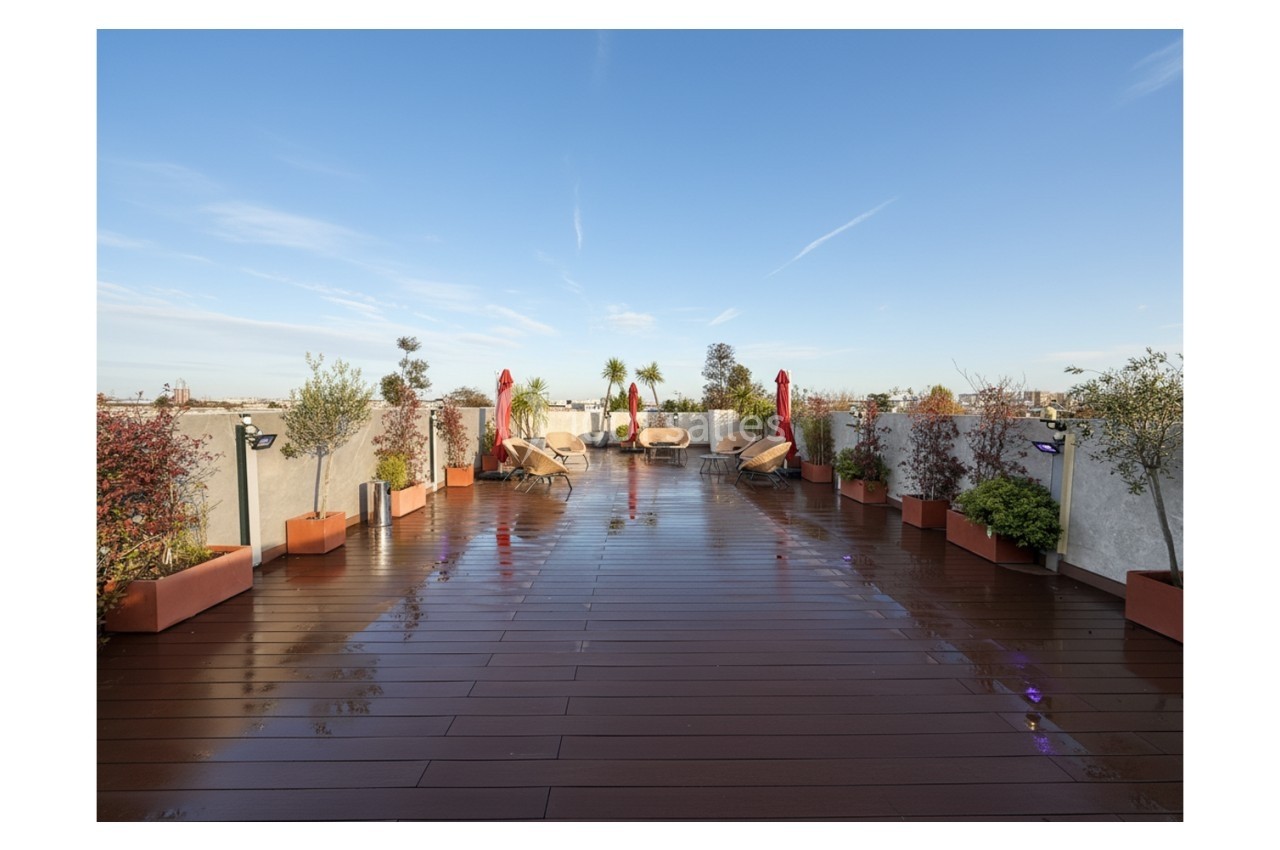Terrasse en bois avec plantes en pots, chaises longues et parasols rouges sous un ciel dégagé.