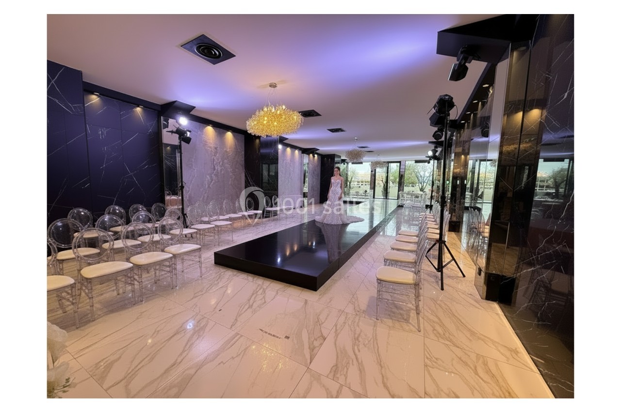Salle élégante avec podium central, chaises transparentes alignées et éclairage tamisé sous un lustre doré.