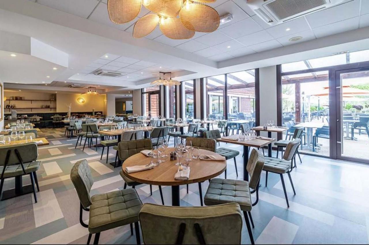 Salle de restaurant lumineuse avec tables dressées, chaises rembourrées et grande baie vitrée donnant sur une terrasse.