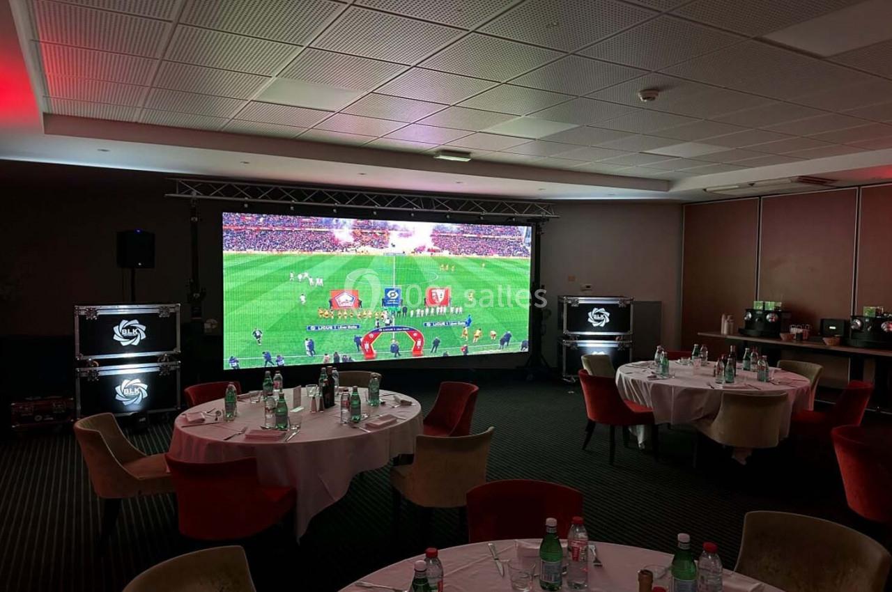 Écran géant diffusant un match de football dans une salle aménagée avec des tables rondes et des chaises.