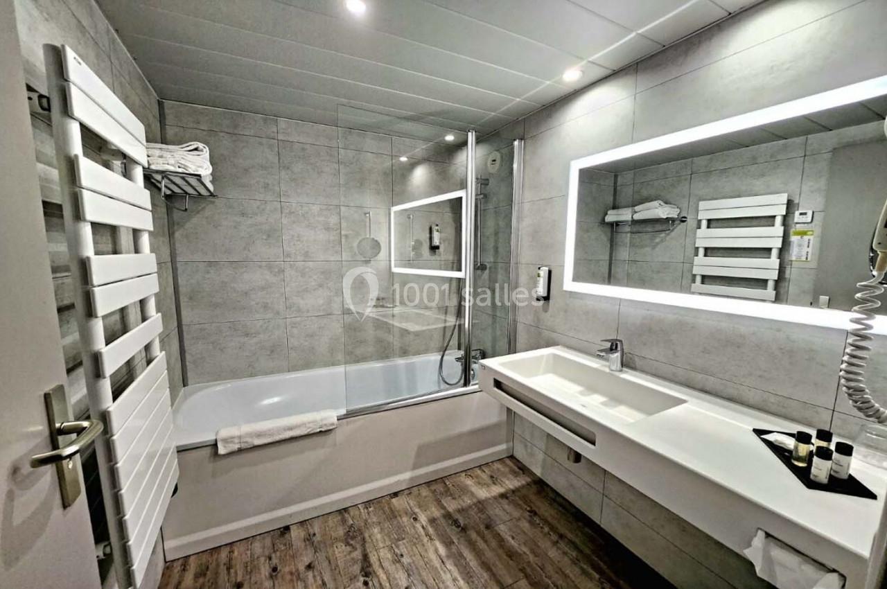 Salle de bain moderne avec baignoire, paroi vitrée, lavabo blanc, miroir éclairé et sol en bois.