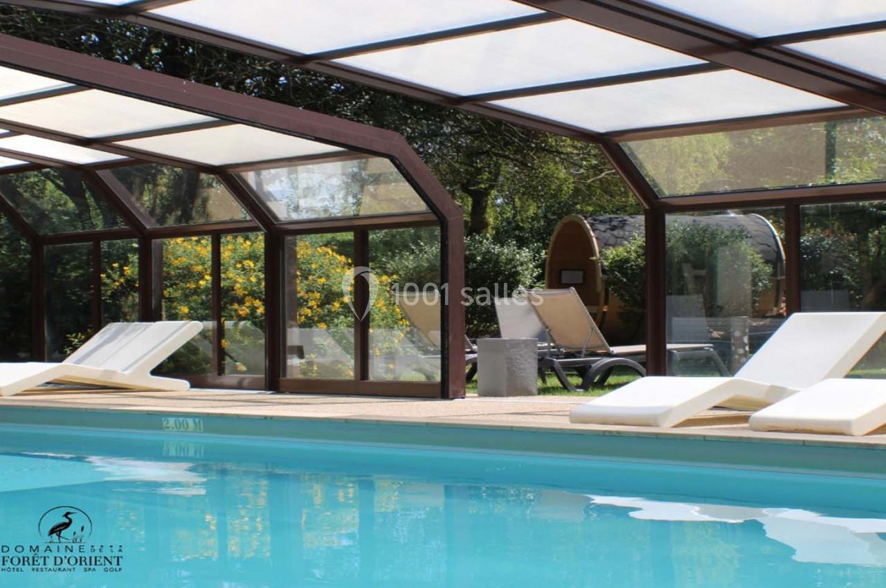 Piscine couverte avec chaises longues blanches, entourée de verdure et de cabanes en bois dans un cadre naturel.