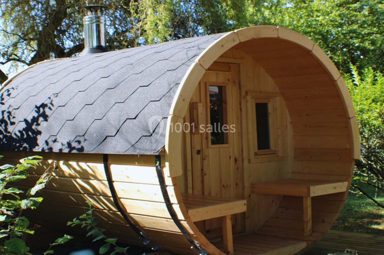 Sauna extérieur en forme de tonneau en bois avec toit sombre, entouré de végétation.