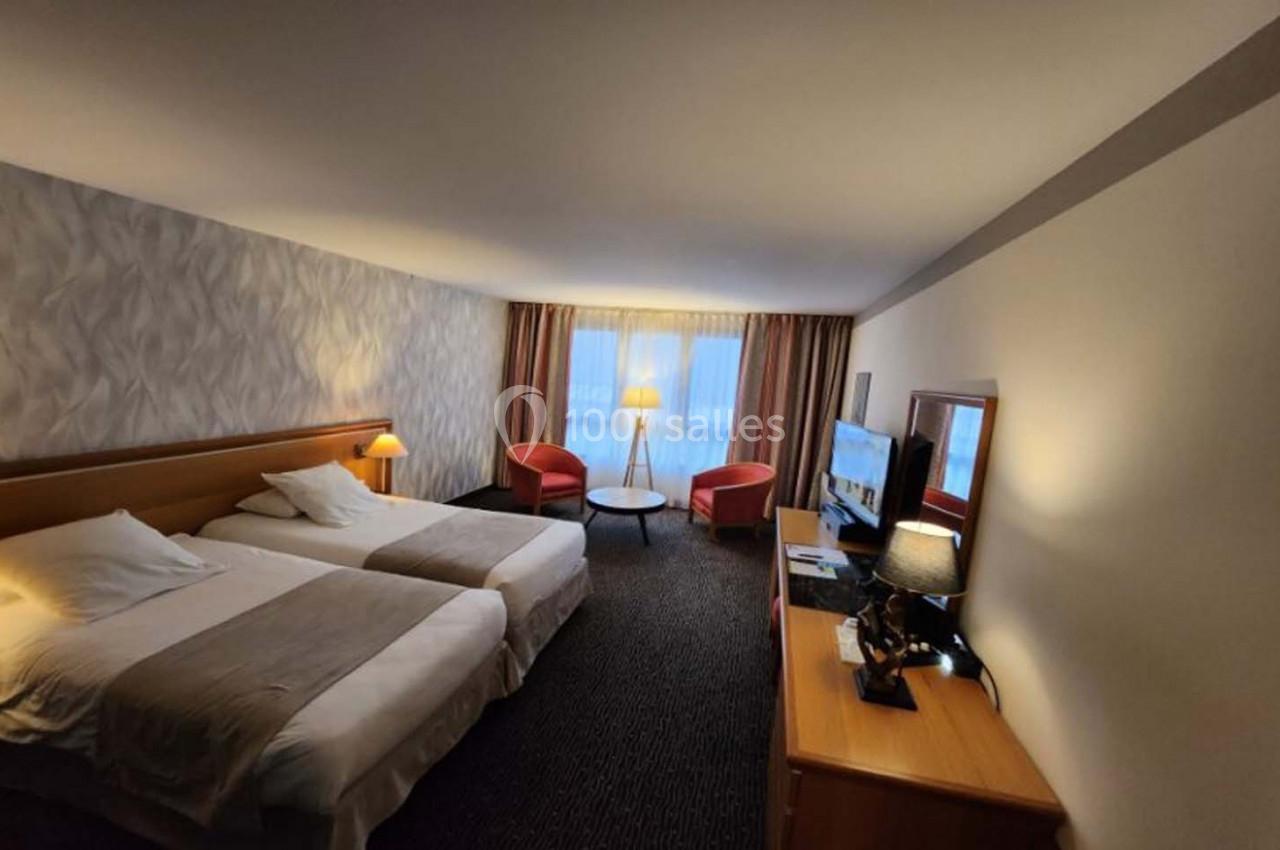 Chambre d'hôtel avec deux lits simples, fauteuils près d'une fenêtre, bureau avec télévision et éclairage chaleureux.