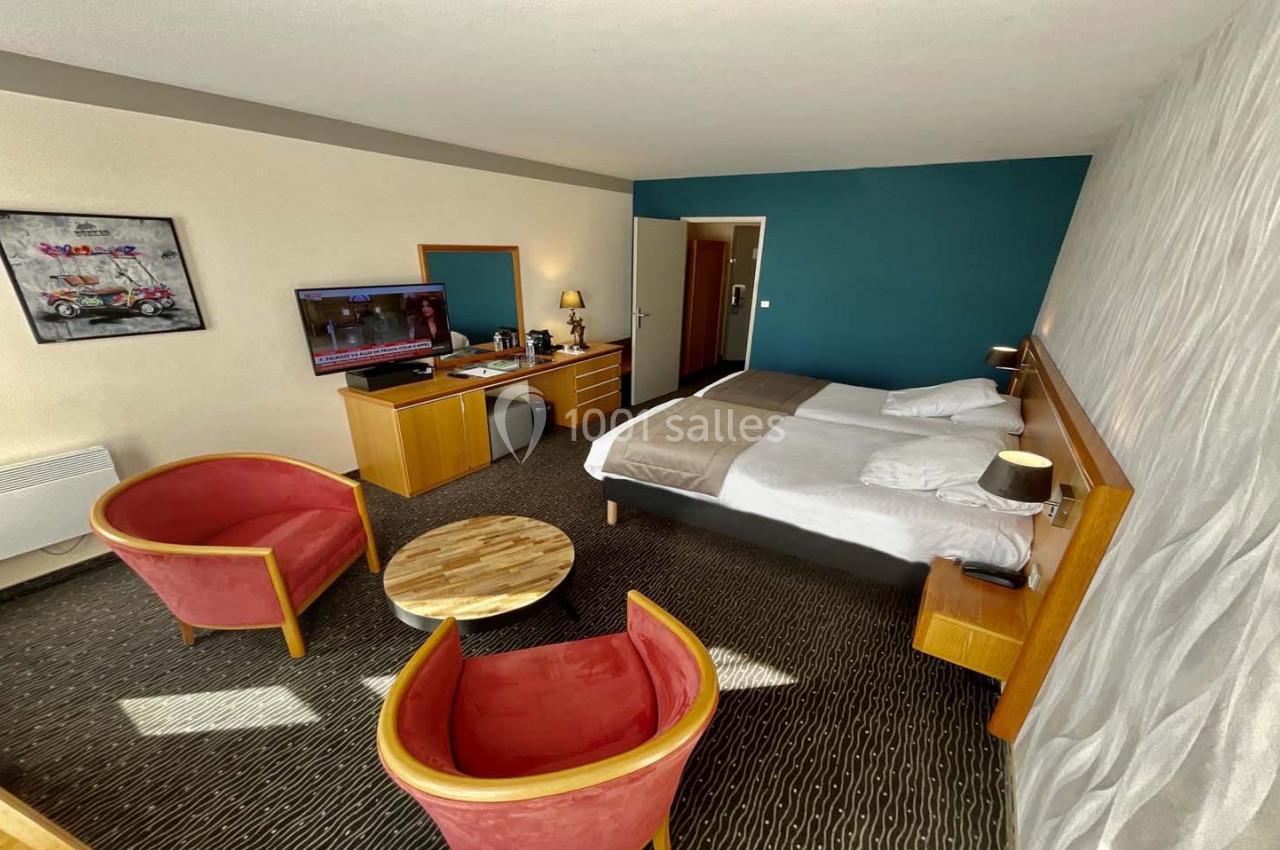 Chambre d'hôtel avec deux lits simples, coin salon avec fauteuils rouges, table basse et télévision allumée.