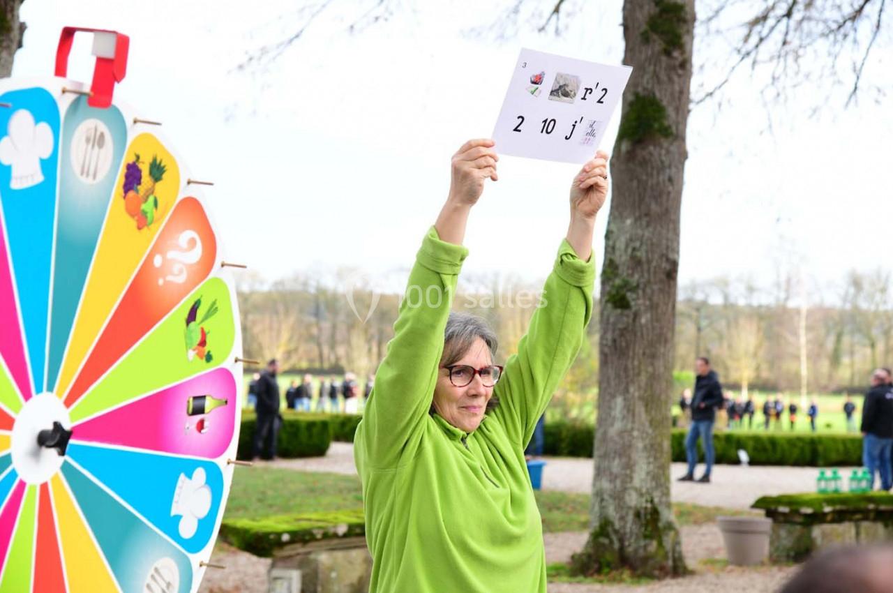 Une personne en pull vert tient une feuille avec des chiffres et des symboles, près d'une roue colorée, dans un parc.