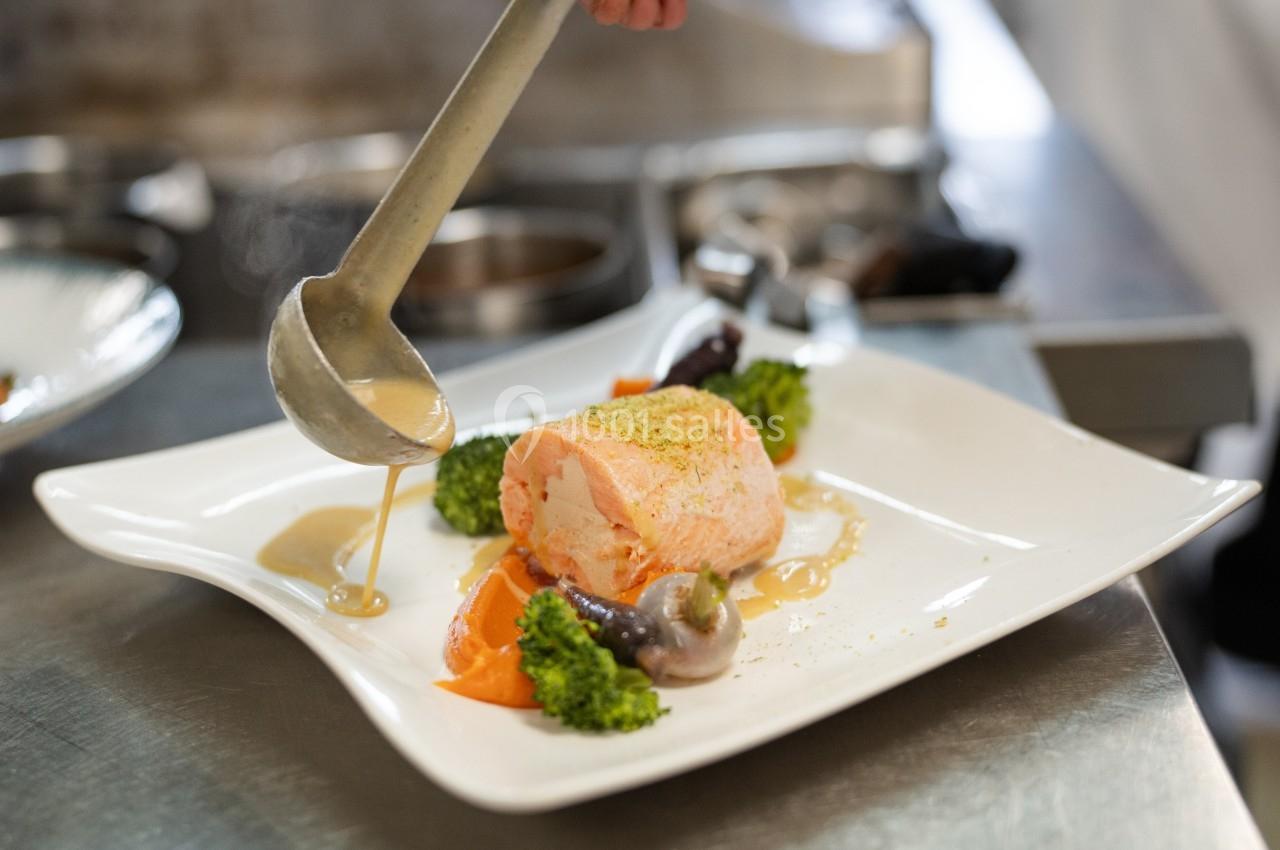 Un filet de saumon servi avec légumes et sauce, dressé sur une assiette blanche dans une cuisine professionnelle.