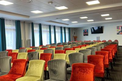 Salle de conférence avec rangées de chaises colorées, un pupitre à l'avant et des panneaux en bois en arrière-plan.