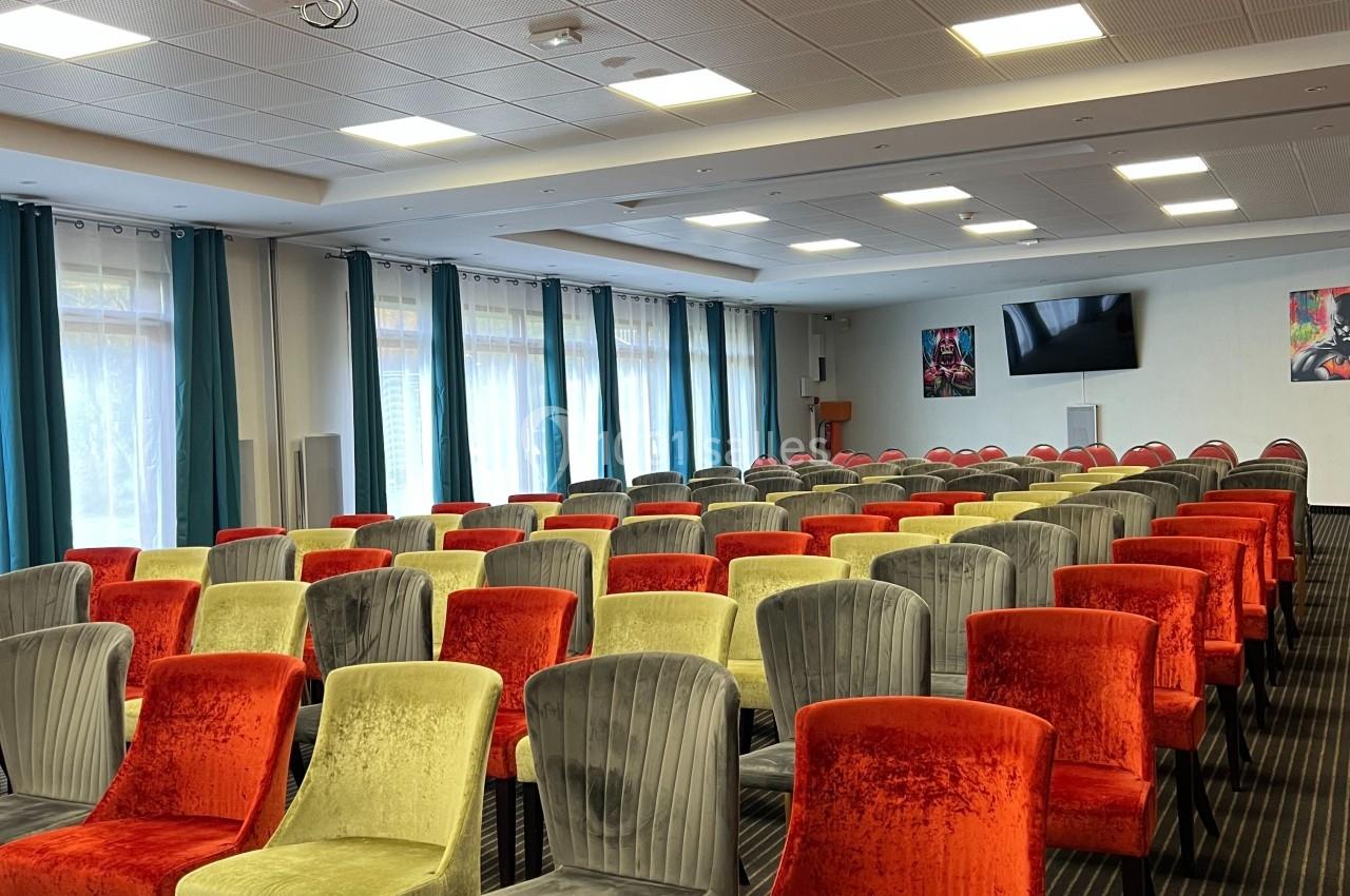 Salle de conférence avec rangées de chaises colorées, plafond à dalles, projecteur et écran mural.