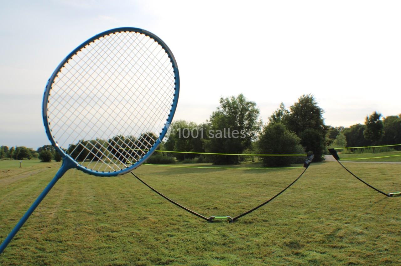 Raquette de badminton bleue tenue près d'un filet installé sur une pelouse dans un parc verdoyant.
