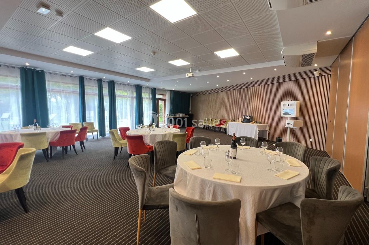 Salle de réception lumineuse avec tables rondes dressées, chaises colorées et grandes baies vitrées donnant sur l'extérieur.