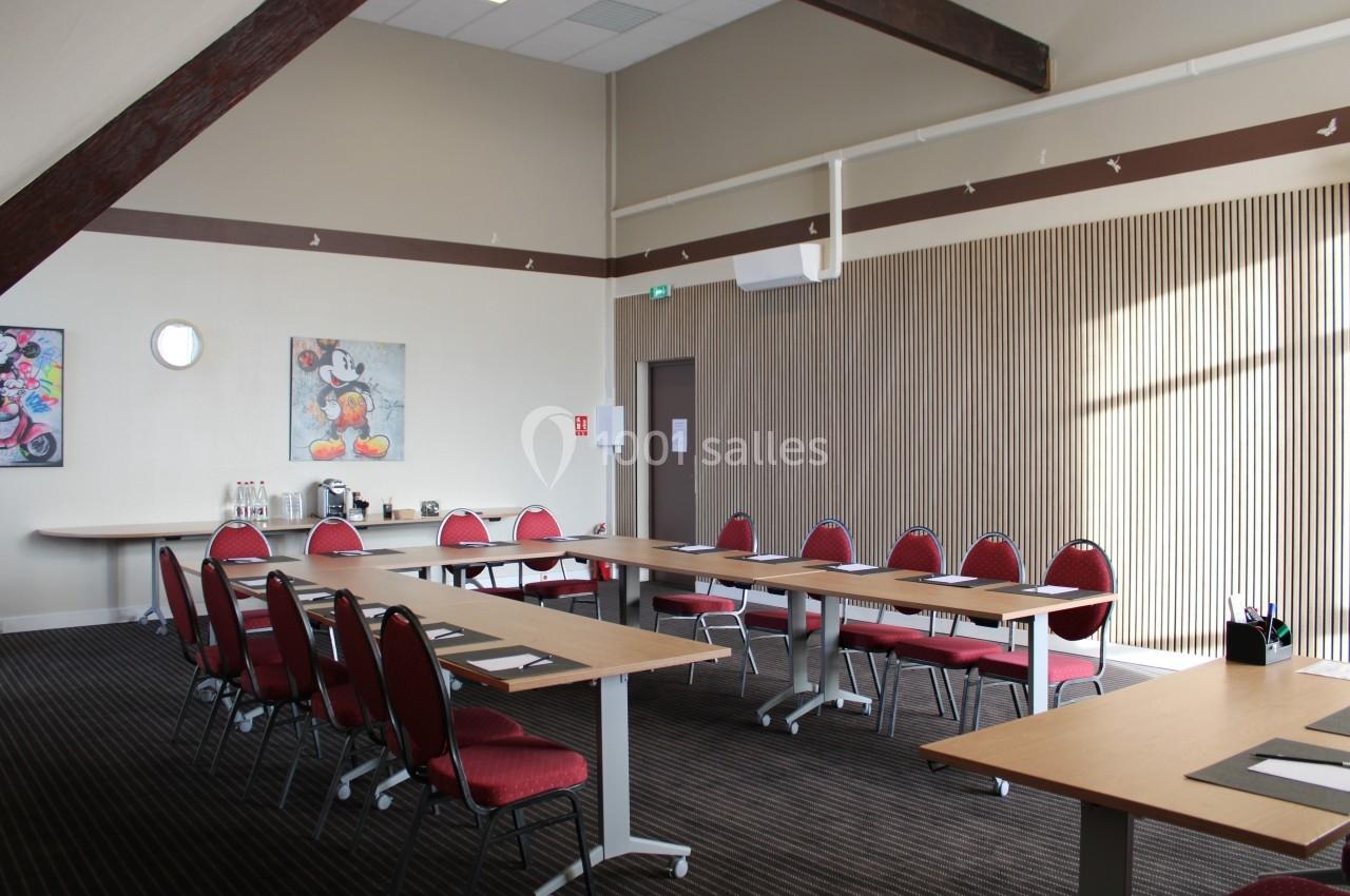 Salle de réunion lumineuse avec tables en U, chaises rouges, tableaux décoratifs et coin café au fond.