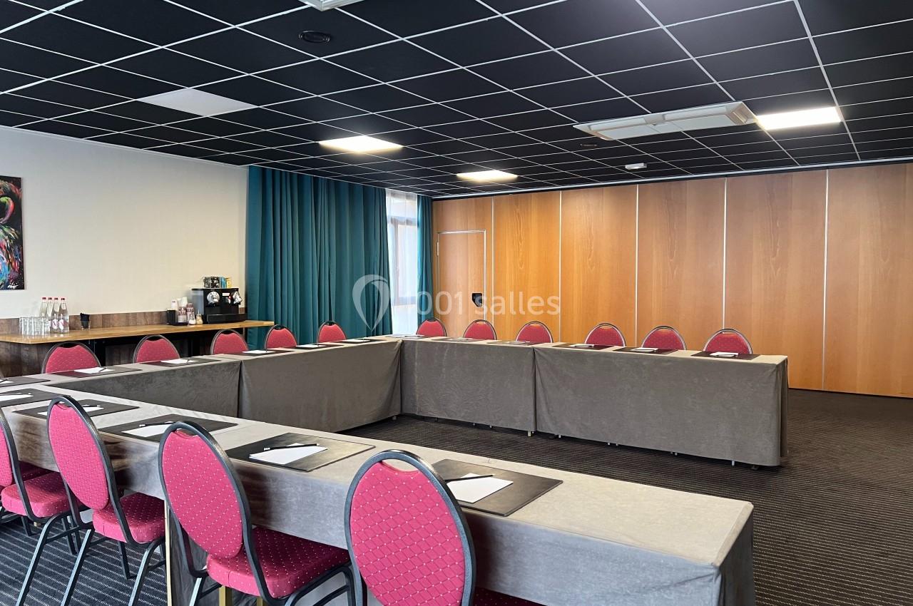 Salle de réunion avec tables disposées en U, chaises rouges, rideaux bleus et coin avec machine à café et bouteilles d'eau.