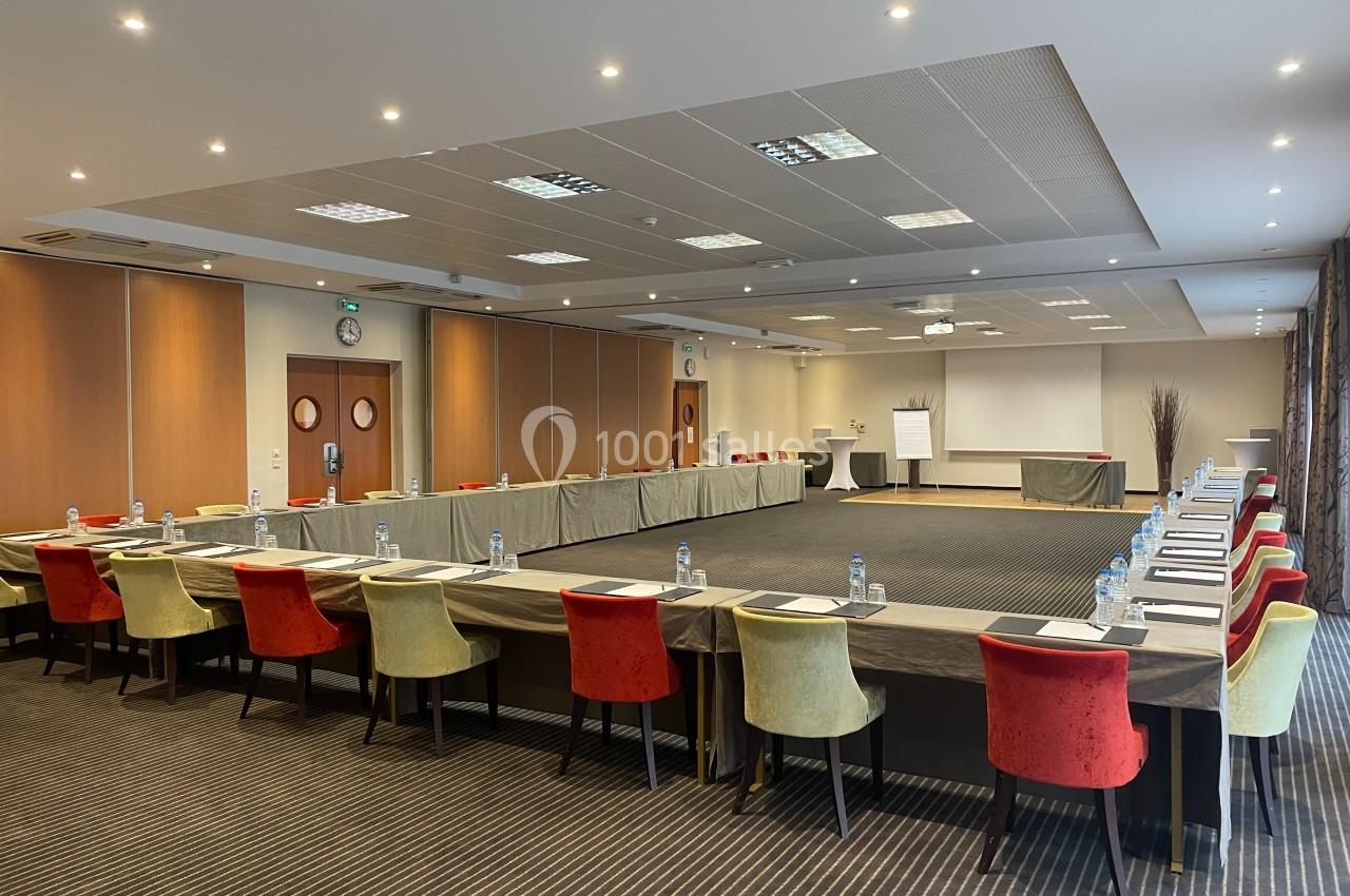 Salle de réunion spacieuse avec tables disposées en U, chaises colorées et équipement de présentation.