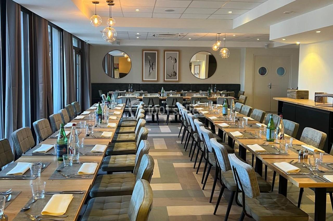 Salle de restaurant lumineuse avec tables alignées, dressées avec nappes blanches, bouteilles d'eau et couverts.