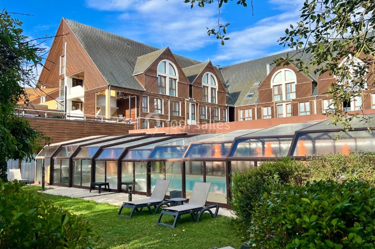 Bâtiment en bois avec grandes fenêtres, piscine couverte et transats dans un jardin verdoyant.