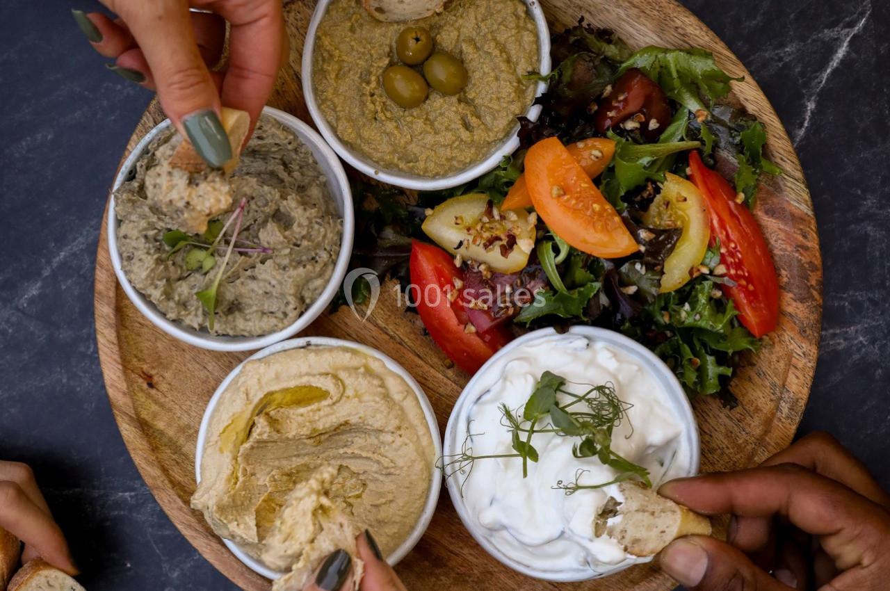 Assiette en bois avec houmous, tzatziki, tapenade, olives et salade, entourée de mains prenant des morceaux de pain.