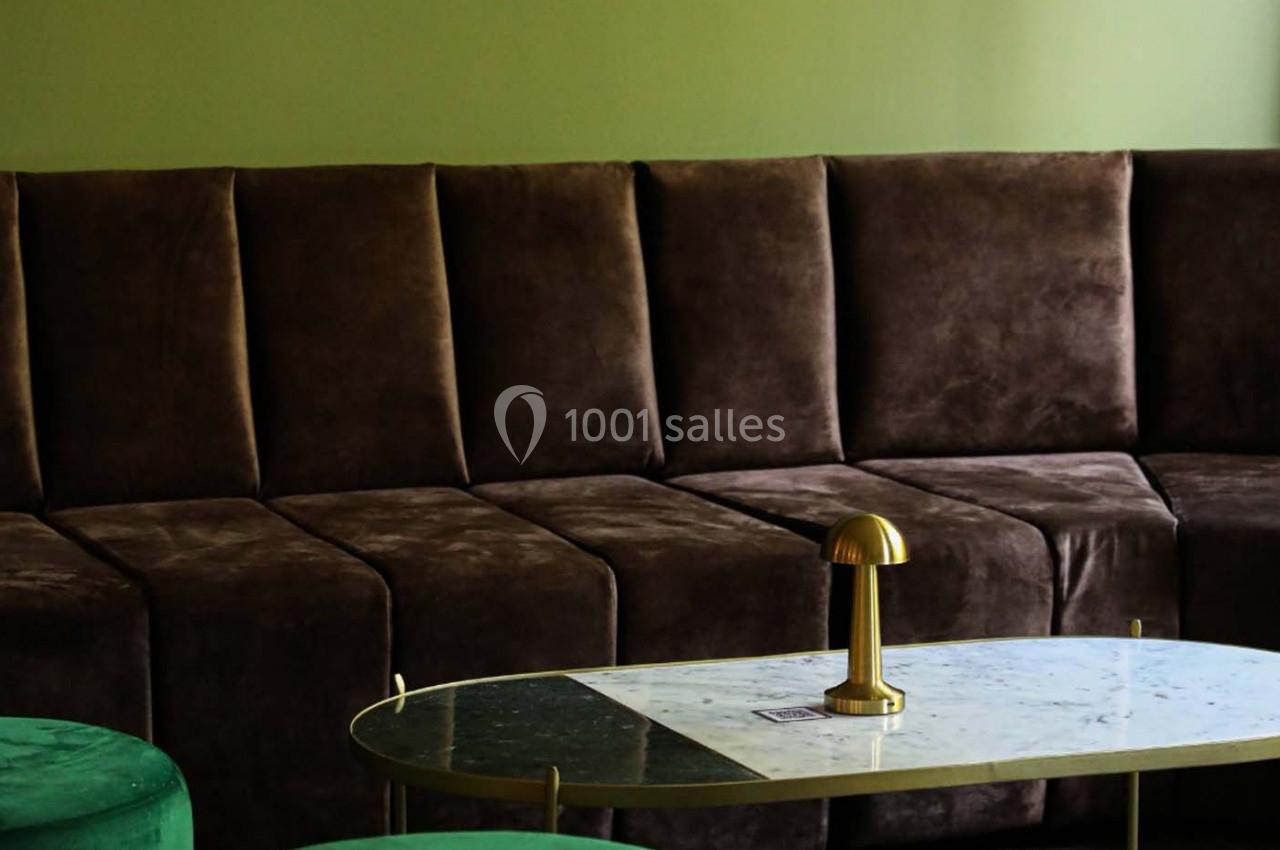 Canapé en velours marron foncé devant une table basse en marbre avec une lampe dorée, dans une pièce aux murs verts.