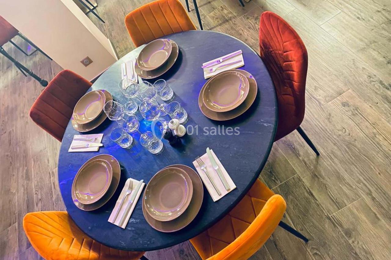 Table ronde dressée avec vaisselle beige, verres et couverts, entourée de chaises orange et rouges sur un sol en bois.