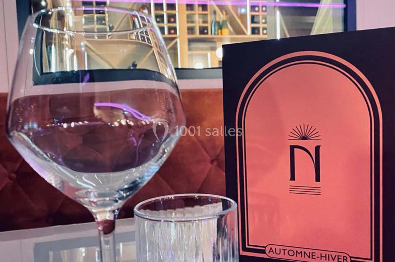 Verre à vin, verre d'eau et menu posé sur une table, avec une étagère à bouteilles en arrière-plan.