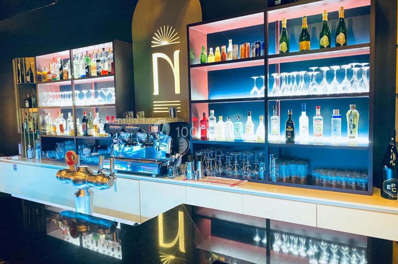 Bar moderne avec étagères éclairées présentant des bouteilles d'alcool, verres et machine à café en arrière-plan.