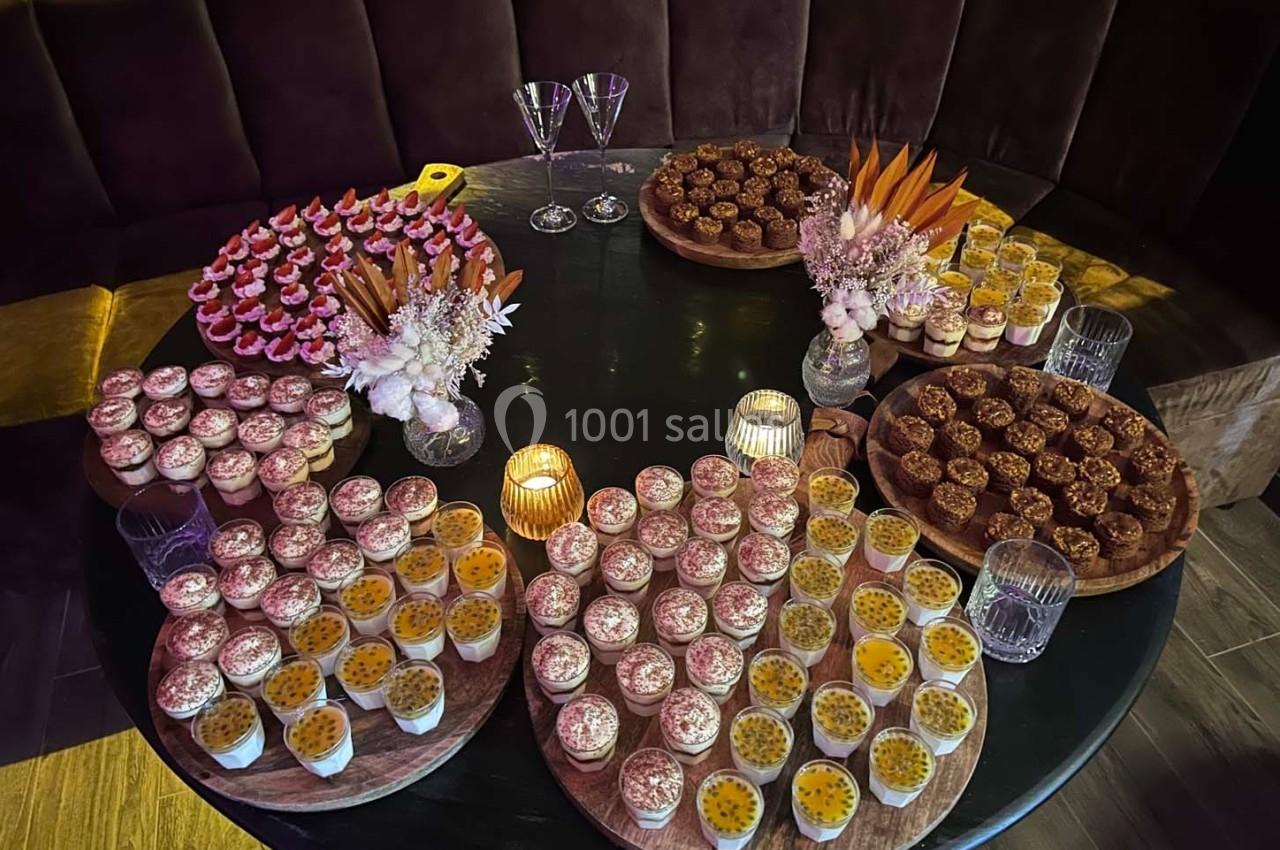 Table ronde garnie de desserts variés, incluant verrines, pâtisseries et décorations florales, éclairée par des bougies.