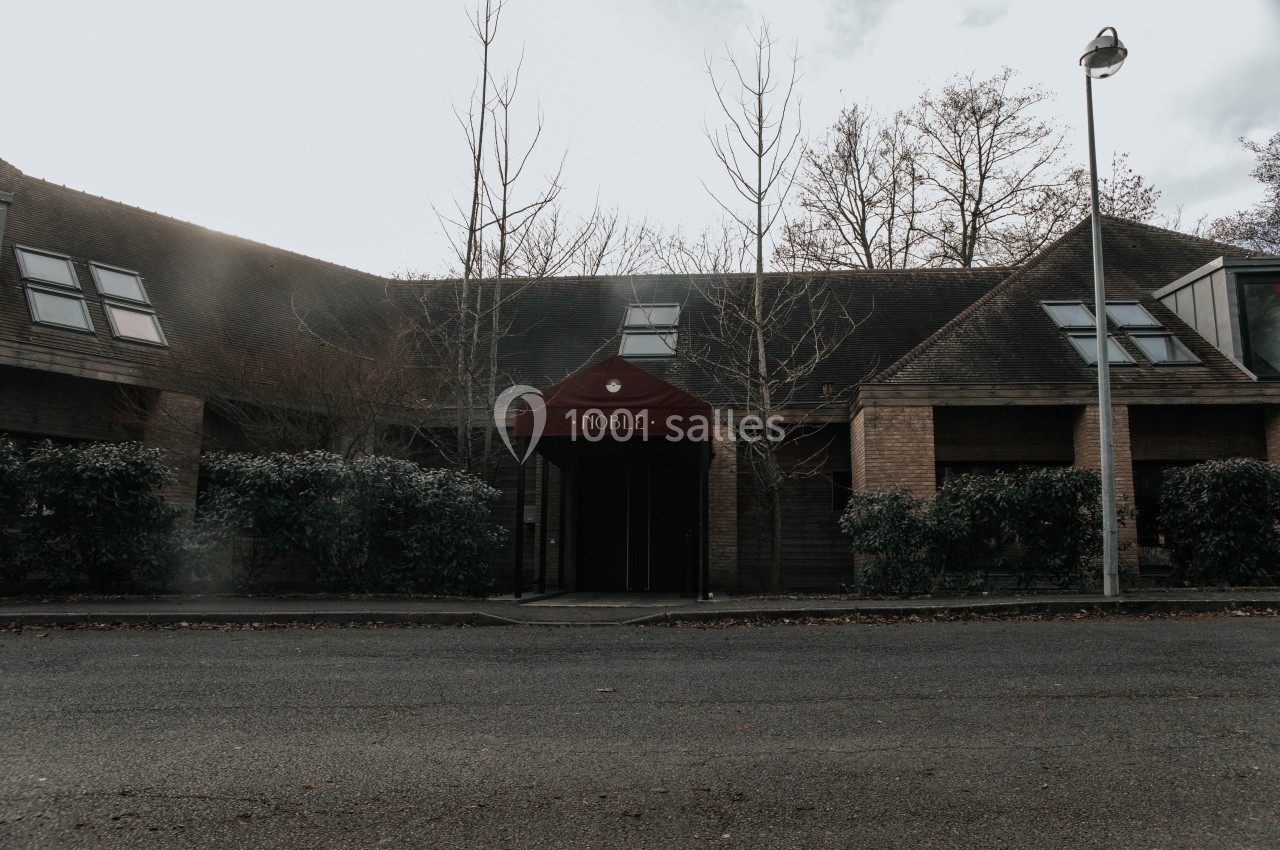 Façade d'un bâtiment en briques avec une entrée couverte par un auvent rouge marqué ’Noble’, entouré de végétation.