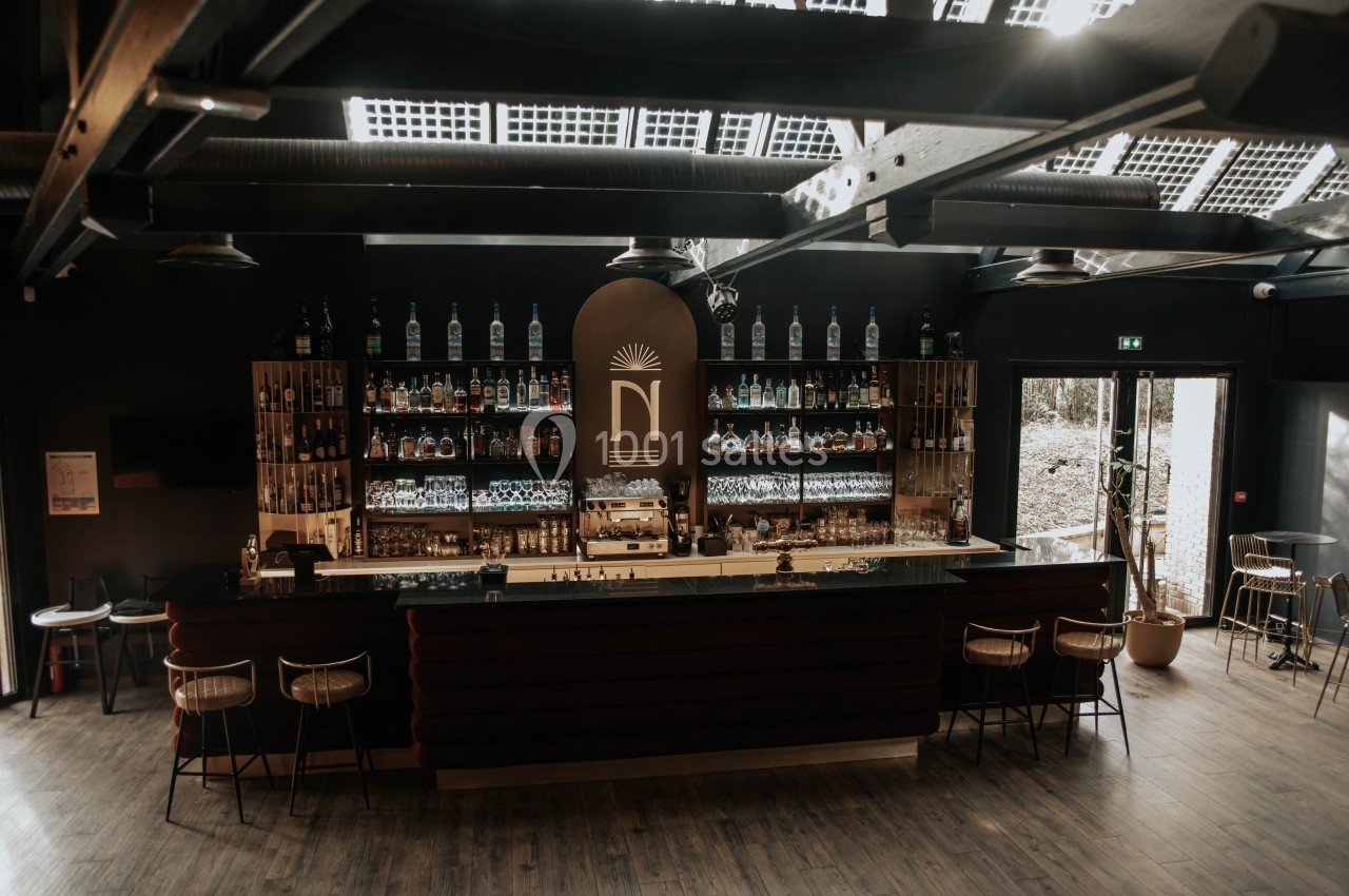 Bar moderne avec comptoir central, étagères remplies de bouteilles et lumière naturelle entrant par le toit vitré.