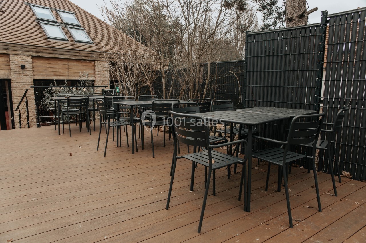 Terrasse en bois avec plusieurs tables et chaises noires, entourée de clôtures et d'arbres dénudés.