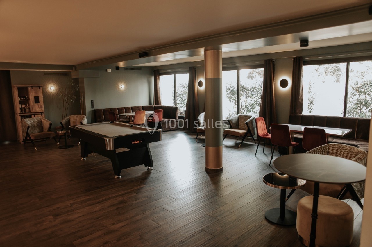 Salle lumineuse avec parquet, billard, tables et chaises, décorée de tons neutres et grandes fenêtres donnant sur l…