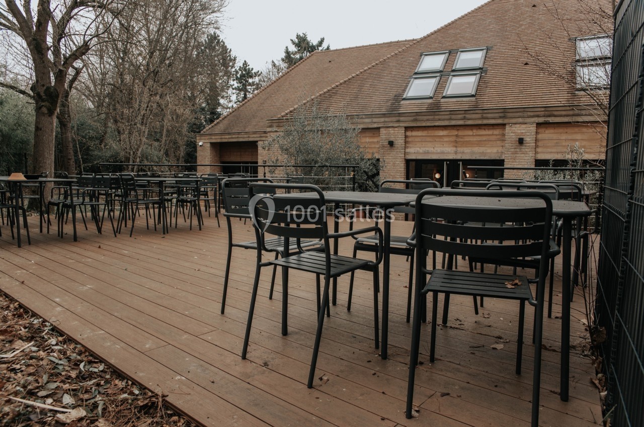Terrasse en bois avec des tables et chaises noires, entourée d'arbres et jouxtant un bâtiment en pierre et bois.