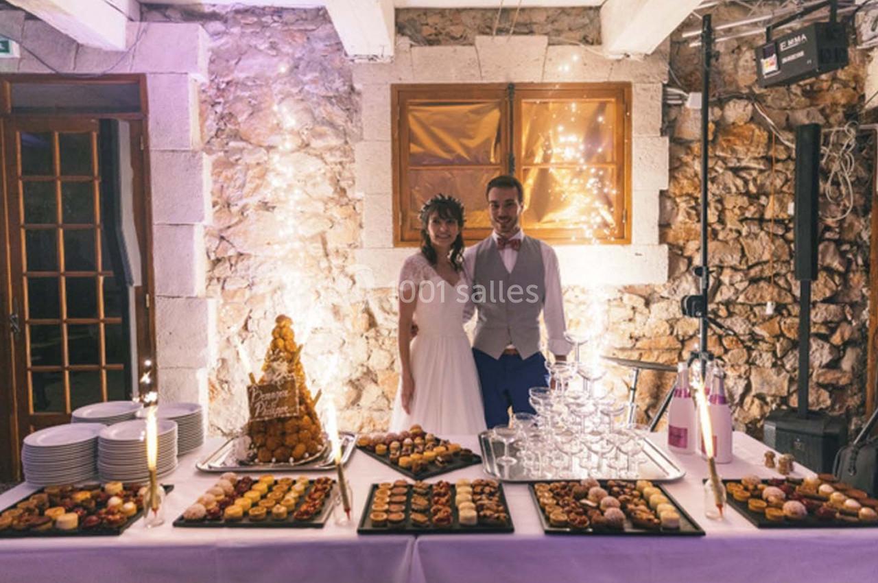 Un couple en tenue de mariage pose devant une table garnie de desserts et d'une pièce montée dans une salle en pierre.