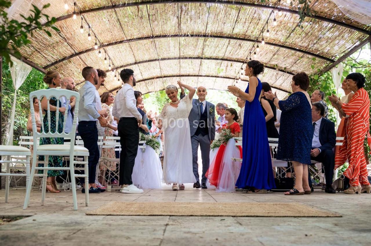 Un couple de mariés marche sous une pergola entouré d'invités applaudissant lors d'une cérémonie en extérieur.