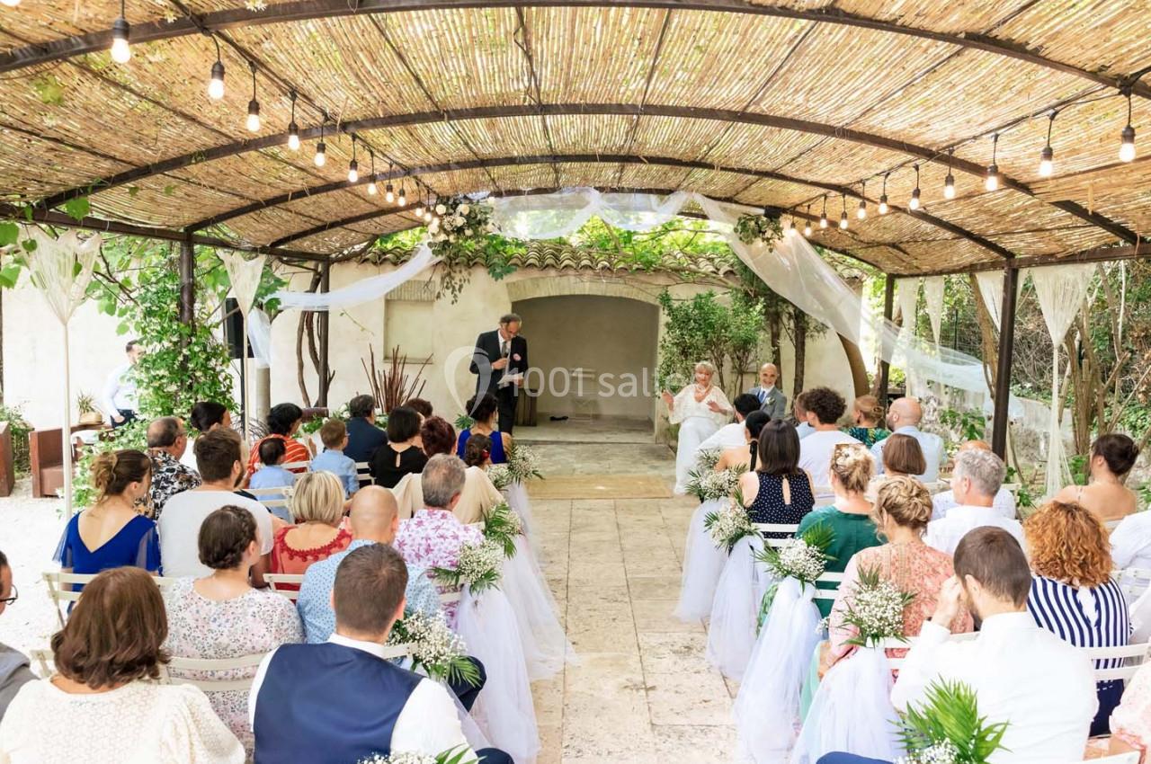 Cérémonie de mariage en plein air sous une pergola décorée, avec des invités assis et un officiant debout.