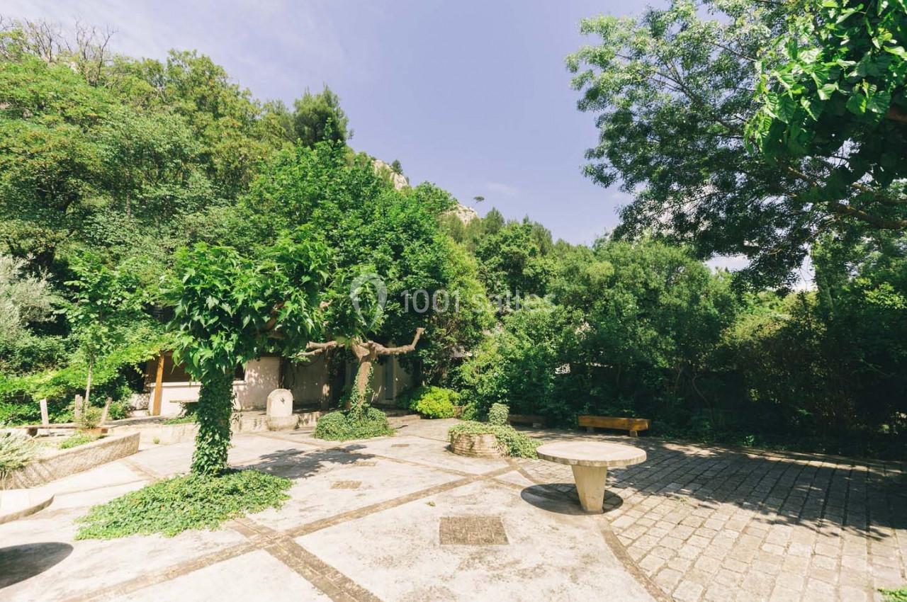Cour extérieure ensoleillée entourée de végétation dense, avec des arbres, des bancs et une table en pierre.